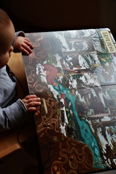 A child is seated and looking at a colorful illustrated book depicting a wintry forest scene with snow, trees, and a flowing river. The illustration includes various animals, such as a red bird and possibly other small creatures. The child is wearing a gray long-sleeve shirt and has their hands resting on the book. The word in Cyrillic at the top edge likely refers to a month or season.