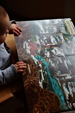 A child is seated and looking at a colorful illustrated book depicting a wintry forest scene with snow, trees, and a flowing river. The illustration includes various animals, such as a red bird and possibly other small creatures. The child is wearing a gray long-sleeve shirt and has their hands resting on the book. The word in Cyrillic at the top edge likely refers to a month or season.