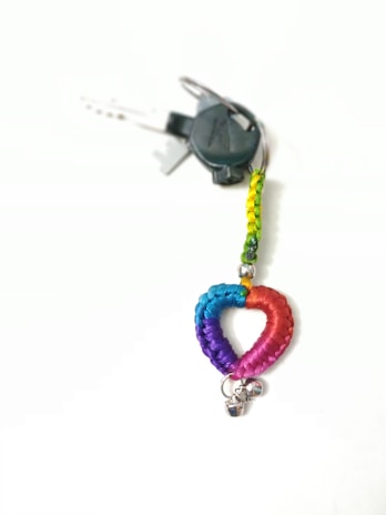 A keychain with a braided heart made of blue, purple, and pink threads is attached to a metal ring with two small silver bells. The braided portion is multicolored, transitioning through shades of blue, purple, and pink. It is connected to a loop of braided thread in yellow and green, from which keys are hanging.