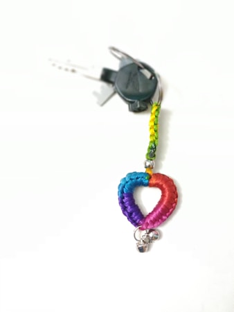 A keychain with a braided heart made of blue, purple, and pink threads is attached to a metal ring with two small silver bells. The braided portion is multicolored, transitioning through shades of blue, purple, and pink. It is connected to a loop of braided thread in yellow and green, from which keys are hanging.