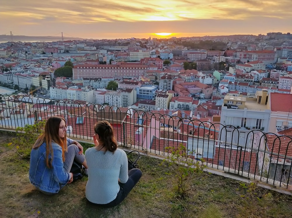 Zwei Frauen sitzen auf Gras in der Nähe eines Geländers mit Blick auf die Stadt unter orangefarbenem Himmel