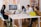 A group of people are seated around a table in a modern office space, engaging in a meeting or collaboration session. The room is decorated with colorful sticky notes on the wall, a large computer monitor displaying a screen, and various laptops and notebooks on the table. The atmosphere is professional yet creative, with a wooden floor and a plant in the foreground.