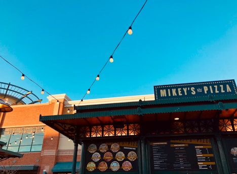 A vibrant blue sky forms the backdrop for an outdoor pizza stand named 'Mikey's Pizza.' The structure features decorative ironwork and a menu display showcasing various pizza options. Festive string lights are suspended above, adding a warm ambiance to the scene.