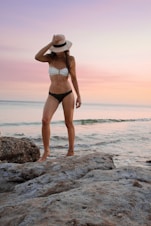 woman in black and white bikini standing on rock formation near sea during golden hour