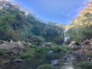 A lush, green landscape features a flowing waterfall in the background, surrounded by dense foliage and trees. Several thatched-roof huts are scattered around, creating a rustic, natural atmosphere. A stream or river runs through the scene, with rocks visible in the water. People are seen walking and playing in the water, enjoying the serene environment.