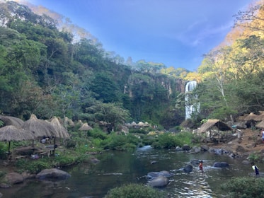 A lush, green landscape features a flowing waterfall in the background, surrounded by dense foliage and trees. Several thatched-roof huts are scattered around, creating a rustic, natural atmosphere. A stream or river runs through the scene, with rocks visible in the water. People are seen walking and playing in the water, enjoying the serene environment.