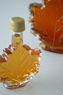 Two glass bottles shaped like maple leaves, filled with amber-colored liquid. One bottle has a gold cap and the liquid appears to be maple syrup. The bottles rest on a smooth, light surface.
