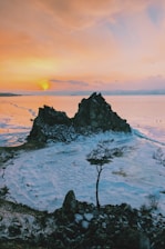 A breathtaking landscape featuring a rugged, rocky formation surrounded by a frozen body of water. The horizon displays a stunning sunset with vibrant hues of orange and pink. A solitary tree stands in the foreground, contrasting with the icy surroundings. Small figures can be seen walking on the ice in the distance.