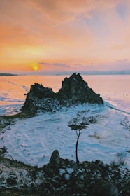 A breathtaking landscape featuring a rugged, rocky formation surrounded by a frozen body of water. The horizon displays a stunning sunset with vibrant hues of orange and pink. A solitary tree stands in the foreground, contrasting with the icy surroundings. Small figures can be seen walking on the ice in the distance.