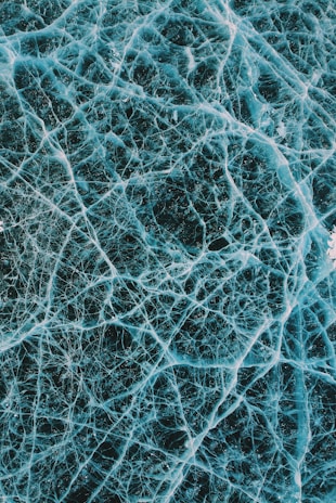 A detailed network of intricate cracks spreads across a surface, resembling cracked ice or glass. The patterns are irregular, forming a complex web of lines in varying thickness.