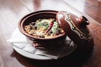 A traditional ceramic dish with intricate patterns contains a baked meal topped with herbs and a red chili pepper. It is placed on a white plate with a paper napkin, on a wooden surface. The lid, decorated with matching designs, is slightly ajar.
