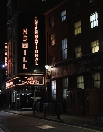 A nighttime urban scene featuring a dimly lit street with neon signage. The signs read 'International' and 'Table Dancing' in bright neon lights. Buildings with multiple illuminated windows line the street. There are traditional red phone booths on the sidewalk and a few street lamps providing additional lighting.