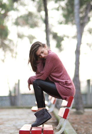 A person wearing a cozy pink sweater, black leggings, and grey boots is sitting sideways on a red and white park bench outdoors. The background features blurred trees and a soft, natural atmosphere.