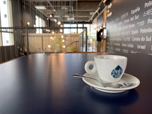 A white ceramic cup with a blue logo sits on a matching saucer with a small spoon placed on a smooth, dark table. The background features a modern, industrial-style cafe interior with hanging lights and a person standing near a counter adorned with international words.