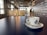 A white ceramic cup with a blue logo sits on a matching saucer with a small spoon placed on a smooth, dark table. The background features a modern, industrial-style cafe interior with hanging lights and a person standing near a counter adorned with international words.
