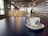 A white ceramic cup with a blue logo sits on a matching saucer with a small spoon placed on a smooth, dark table. The background features a modern, industrial-style cafe interior with hanging lights and a person standing near a counter adorned with international words.