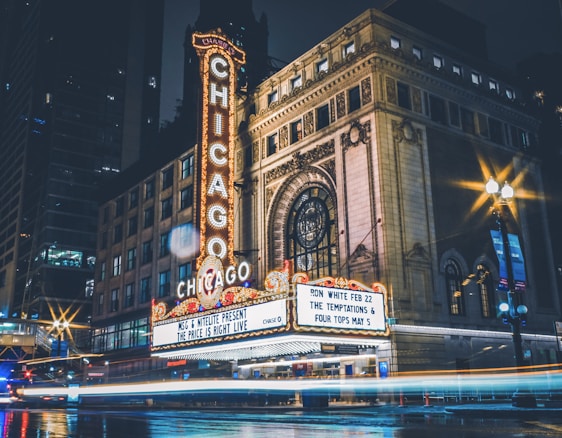 A theater sign lit up in bright neon lights with the word 'Chicago' prominently displayed. The marquee below announces upcoming shows and events. The building has an ornate facade with large windows and decorative architectural elements. The scene is set at night, with light trails from passing vehicles and reflections on the wet pavement.