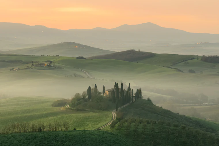 Mist layered over the Val d'Orcia at dawn, a cypress-ringed farmhouse on the slope