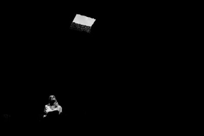 A person is seated in a dimly lit space with a shaft of light coming from a rectangular opening above. The person appears contemplative, gazing upwards towards the light source.
