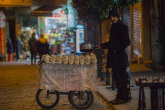 A street vendor, dressed warmly in dark clothing with a beanie, stands beside a cart adorned with a floral cloth. The cart has several bags of popcorn or roasted chestnuts displayed on top. The setting is a dimly lit street with cobblestones, likely in an evening or night-time urban environment. In the background, there is a brightly lit shop with blurry figures of people walking by.