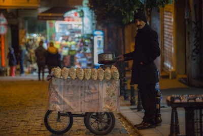 A street vendor, dressed warmly in dark clothing with a beanie, stands beside a cart adorned with a floral cloth. The cart has several bags of popcorn or roasted chestnuts displayed on top. The setting is a dimly lit street with cobblestones, likely in an evening or night-time urban environment. In the background, there is a brightly lit shop with blurry figures of people walking by.
