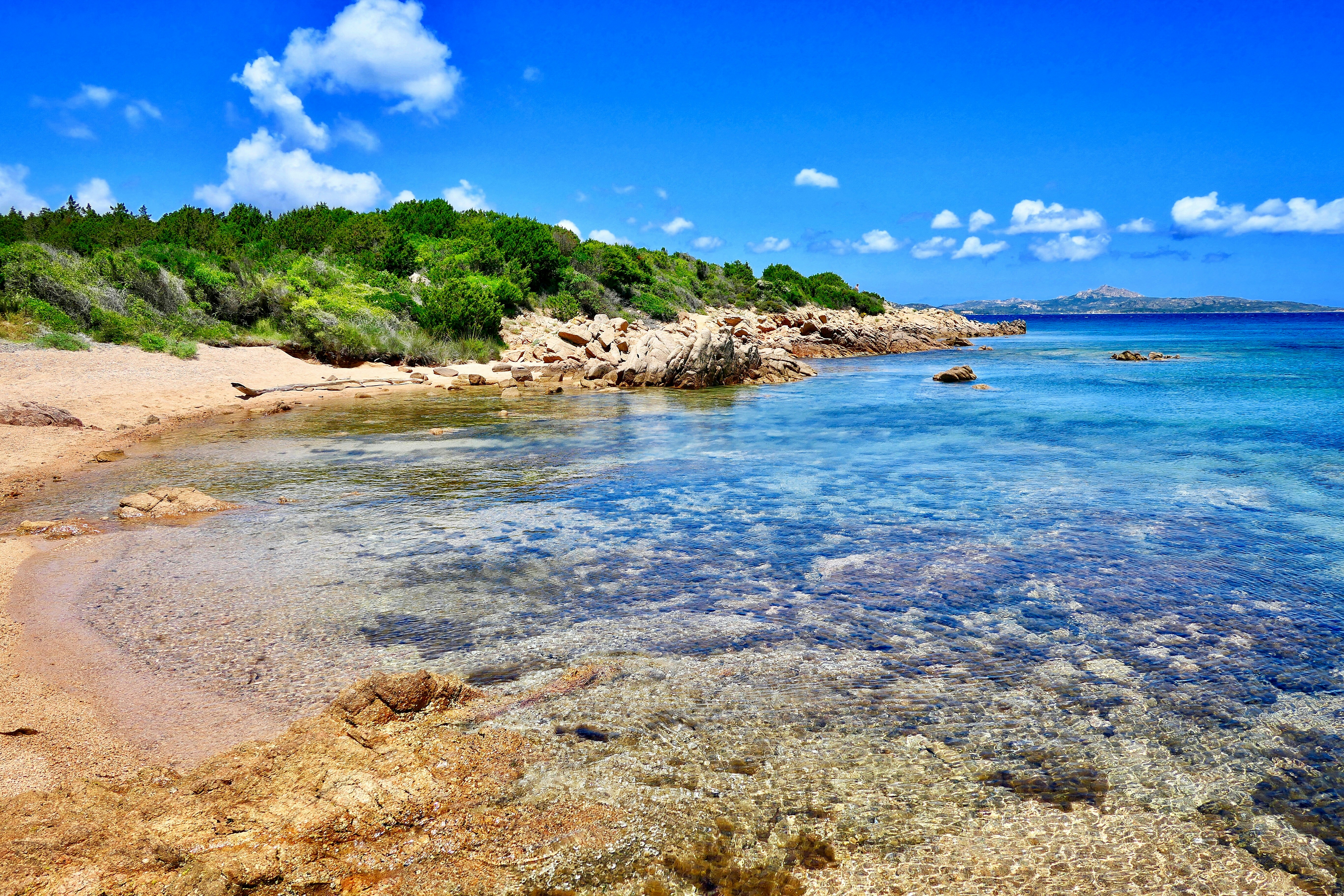 a beach with clear blue water surrounded by trees, Spiaggia Li Piscini, Palau, Sardinia, Italy