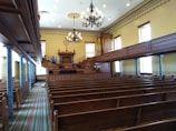 An interior of a vintage courtroom or assembly hall featuring rows of wooden benches, ornate chandeliers hanging from the ceiling, a judge's podium with an organ behind it, and blue marble columns lining the aisle. The walls are painted a soft yellow, and there are several tall windows letting in natural light.