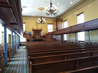 An interior of a vintage courtroom or assembly hall featuring rows of wooden benches, ornate chandeliers hanging from the ceiling, a judge's podium with an organ behind it, and blue marble columns lining the aisle. The walls are painted a soft yellow, and there are several tall windows letting in natural light.