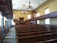 An interior of a vintage courtroom or assembly hall featuring rows of wooden benches, ornate chandeliers hanging from the ceiling, a judge's podium with an organ behind it, and blue marble columns lining the aisle. The walls are painted a soft yellow, and there are several tall windows letting in natural light.