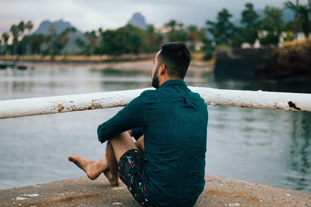 man sitting near metal bar in front of body of water