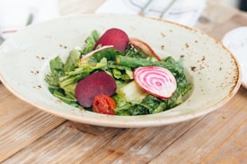 A ceramic plate contains a vibrant salad consisting of fresh greens, slices of red beets, a striped beet slice, and a small red cherry tomato. The plate has a speckled pattern and rests on a wooden surface, adding a rustic touch to the presentation. The ingredients are arranged aesthetically, highlighting their contrasting colors and textures.