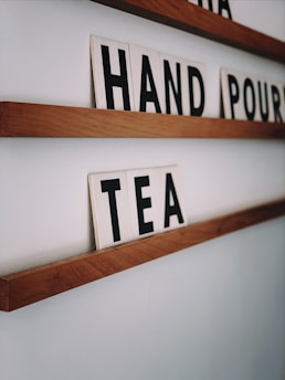 Wooden shelves mounted on a white wall hold cards with black letters. The visible words formed are 'HAND', 'POUR', and 'TEA'. The shelves have a minimalistic design, emphasizing simplicity and functionality.