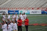 A group of athletes are gathered on a sports field wearing white jerseys with red lettering and holding lacrosse sticks. They appear to be preparing for or concluding a game. The background shows a large number of empty bleachers and various advertisements on a surrounding wall.