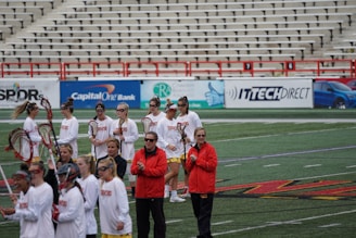 A group of athletes are gathered on a sports field wearing white jerseys with red lettering and holding lacrosse sticks. They appear to be preparing for or concluding a game. The background shows a large number of empty bleachers and various advertisements on a surrounding wall.