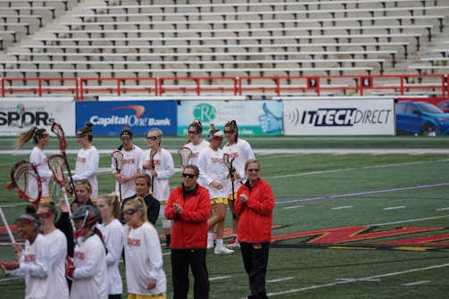 A group of athletes are gathered on a sports field wearing white jerseys with red lettering and holding lacrosse sticks. They appear to be preparing for or concluding a game. The background shows a large number of empty bleachers and various advertisements on a surrounding wall.