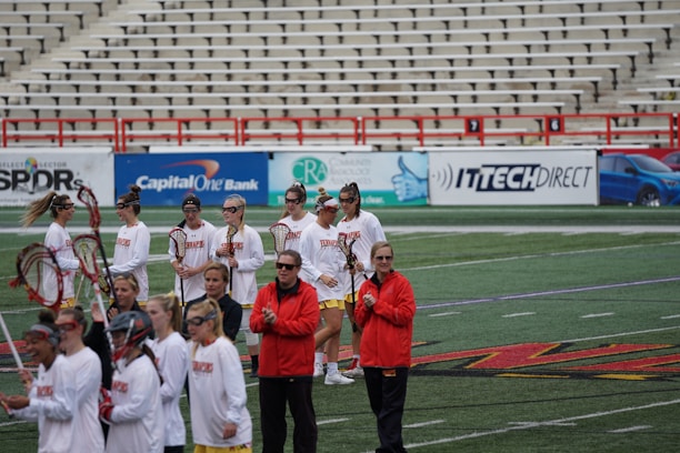 A group of athletes are gathered on a sports field wearing white jerseys with red lettering and holding lacrosse sticks. They appear to be preparing for or concluding a game. The background shows a large number of empty bleachers and various advertisements on a surrounding wall.