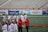 A group of athletes are gathered on a sports field wearing white jerseys with red lettering and holding lacrosse sticks. They appear to be preparing for or concluding a game. The background shows a large number of empty bleachers and various advertisements on a surrounding wall.
