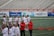 A group of athletes are gathered on a sports field wearing white jerseys with red lettering and holding lacrosse sticks. They appear to be preparing for or concluding a game. The background shows a large number of empty bleachers and various advertisements on a surrounding wall.