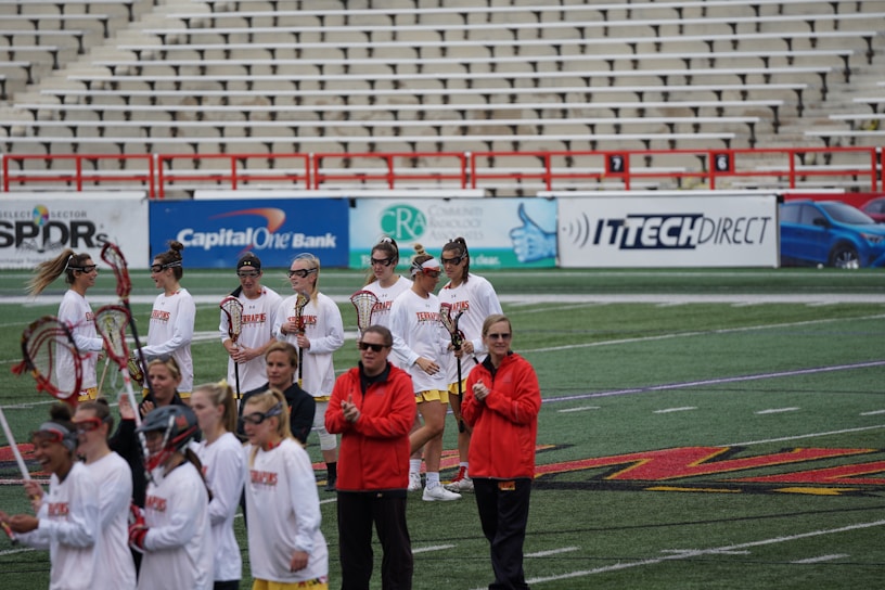 A group of athletes are gathered on a sports field wearing white jerseys with red lettering and holding lacrosse sticks. They appear to be preparing for or concluding a game. The background shows a large number of empty bleachers and various advertisements on a surrounding wall.