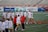 A group of female lacrosse players and coaches are gathered on a sports field. The women are wearing white jerseys with the word 'Terrapins' and holding lacrosse sticks. The scene appears to be during a practice or warm-up session. Behind them are sports advertisements, and the setting is an outdoor stadium.