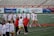 A group of female lacrosse players and coaches are gathered on a sports field. The women are wearing white jerseys with the word 'Terrapins' and holding lacrosse sticks. The scene appears to be during a practice or warm-up session. Behind them are sports advertisements, and the setting is an outdoor stadium.