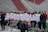 A group of female athletes in white jerseys and yellow shorts are standing in a line. They appear to be on a sports field with empty red and white stadium seats in the background. Several individuals are wearing helmets. Athletic equipment and chairs are in the foreground.