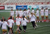 A group of female lacrosse players in matching uniforms stand in a stadium, some holding lacrosse sticks and others engaged in conversation or celebration. They are on a sports field with advertisements visible in the background, and the setting conveys a team environment.