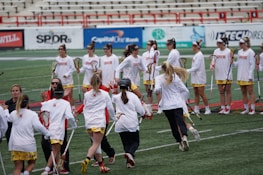 A group of female lacrosse players in matching uniforms stand in a stadium, some holding lacrosse sticks and others engaged in conversation or celebration. They are on a sports field with advertisements visible in the background, and the setting conveys a team environment.