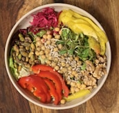 A vibrant salad bowl featuring a variety of fresh ingredients including chickpeas, pistachios, walnuts, sunflower seeds, sliced red peppers, yellow peppers, greens, and a portion of red pickled cabbage. These colorful ingredients are arranged artfully, creating a visually appealing and nutritious dish.