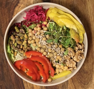 A vibrant salad bowl featuring a variety of fresh ingredients including chickpeas, pistachios, walnuts, sunflower seeds, sliced red peppers, yellow peppers, greens, and a portion of red pickled cabbage. These colorful ingredients are arranged artfully, creating a visually appealing and nutritious dish.