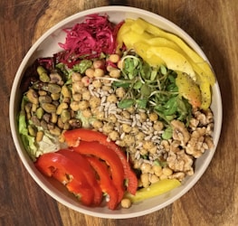 A vibrant salad bowl featuring a variety of fresh ingredients including chickpeas, pistachios, walnuts, sunflower seeds, sliced red peppers, yellow peppers, greens, and a portion of red pickled cabbage. These colorful ingredients are arranged artfully, creating a visually appealing and nutritious dish.
