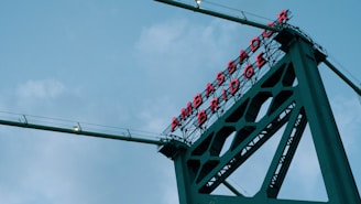 A towering section of a bridge with 'AMBASSADOR BRIDGE' written in red neon letters. The bridge's structure is visible against a backdrop of a cloudy sky, showcasing metal beams and cables.