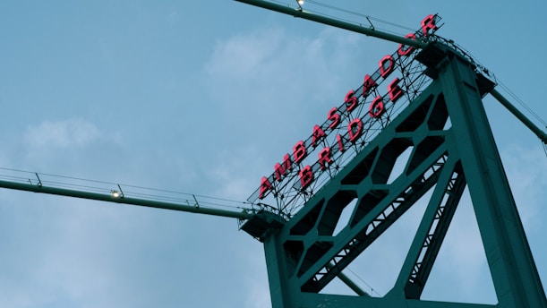 A towering section of a bridge with 'AMBASSADOR BRIDGE' written in red neon letters. The bridge's structure is visible against a backdrop of a cloudy sky, showcasing metal beams and cables.