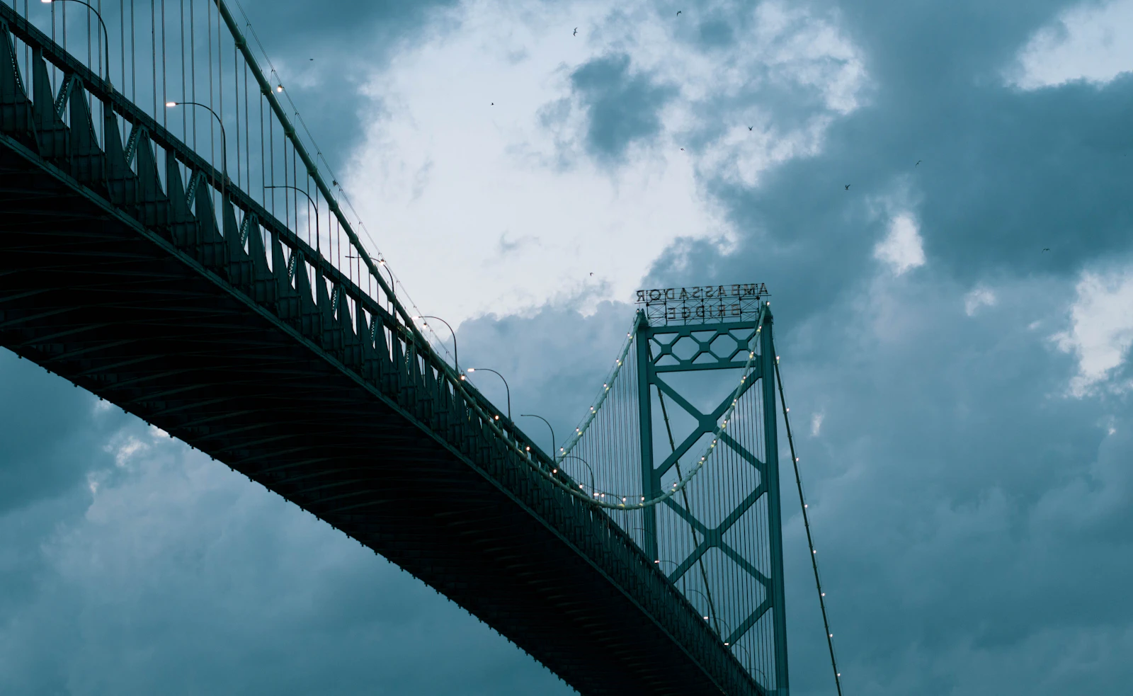 A steel suspension bridge seen from below, its tower set against a dark, cloudy blue sky.